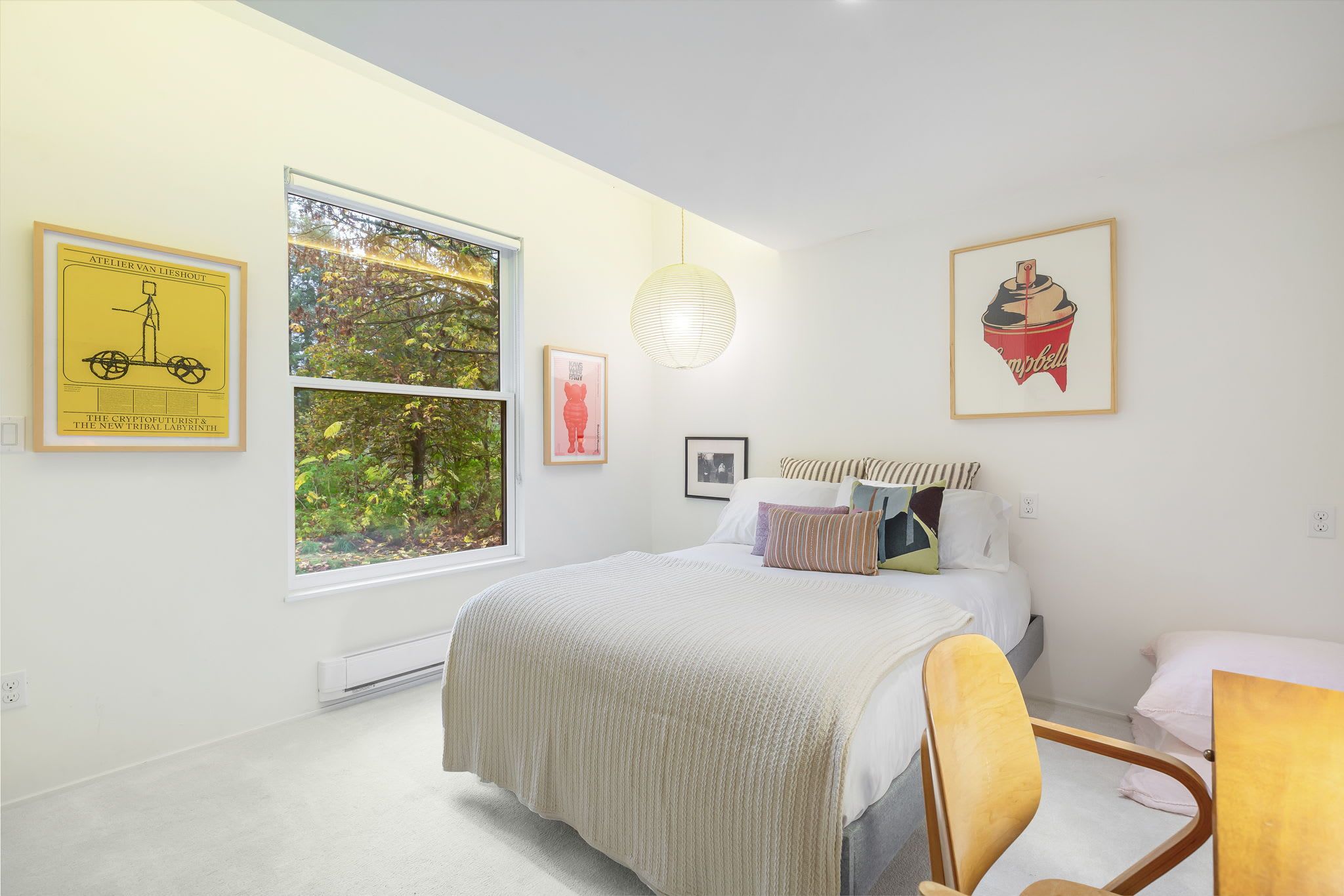 Une chambre avec une grande fenêtre, un lit avec de nombreux oreillers, une chaise et des œuvres d’art murales, notamment un tableau encadré représentant un vélo et un cupcake.