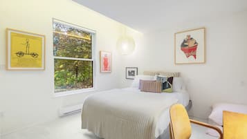 Quarto com uma janela grande, uma cama com várias almofadas, uma cadeira e decoração na parede, incluindo uma fotografia emoldurada de uma bicicleta e um cupcake.