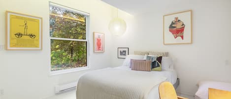Une chambre avec une grande fenêtre, un lit avec de nombreux oreillers, une chaise et des œuvres d’art murales, notamment un tableau encadré représentant un vélo et un cupcake.