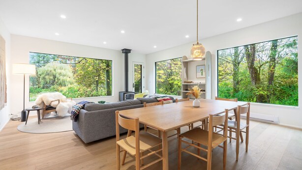 A modern living room with a wooden dining table, grey sofa, and large windows offering a view of greenery. - Sleeps 6 · 3BR Craftsman · Mountain Views · Near Skagit River (Concrete)