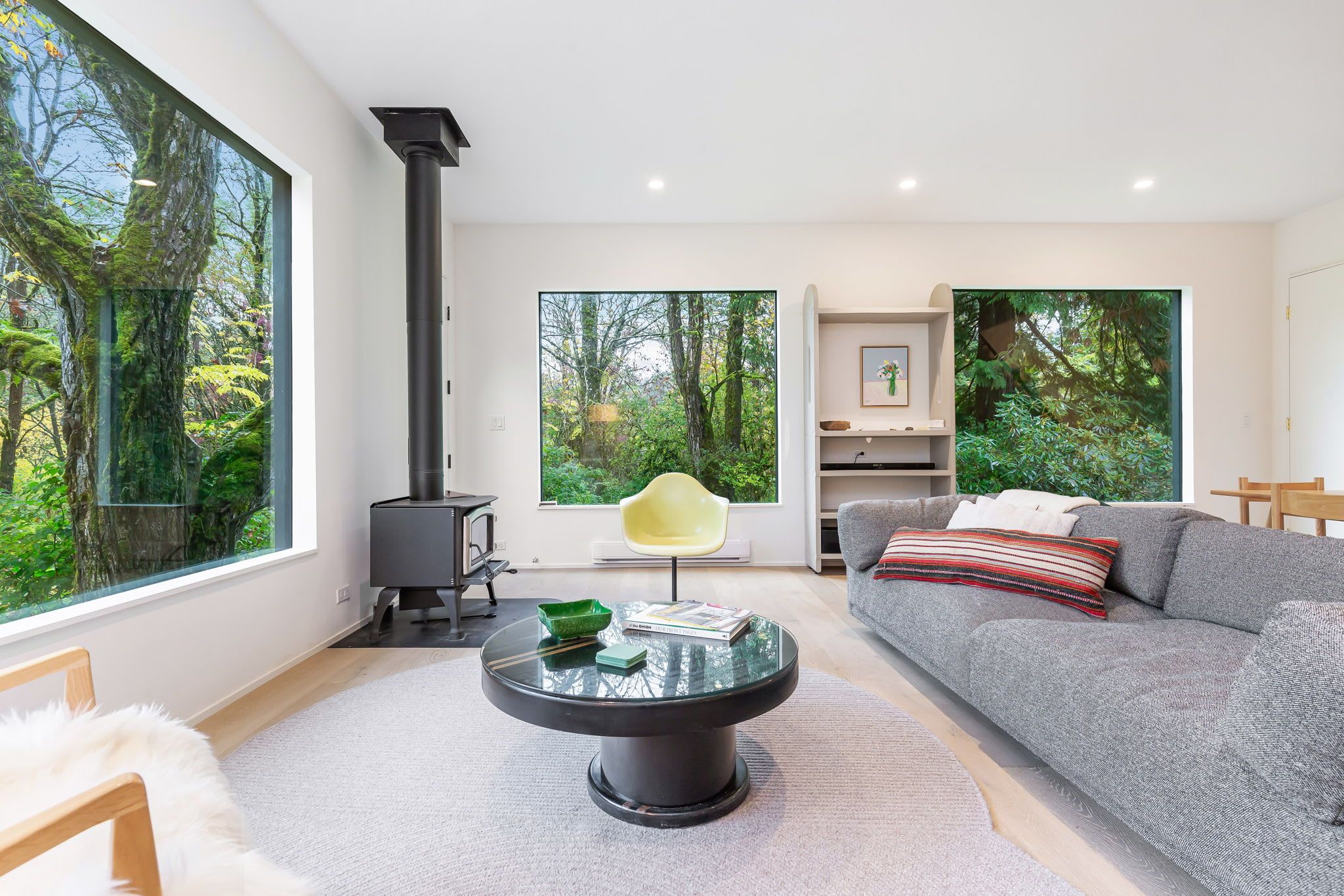 A modern living room with a large window, a round glass coffee table, a grey sofa, and a black fireplace.