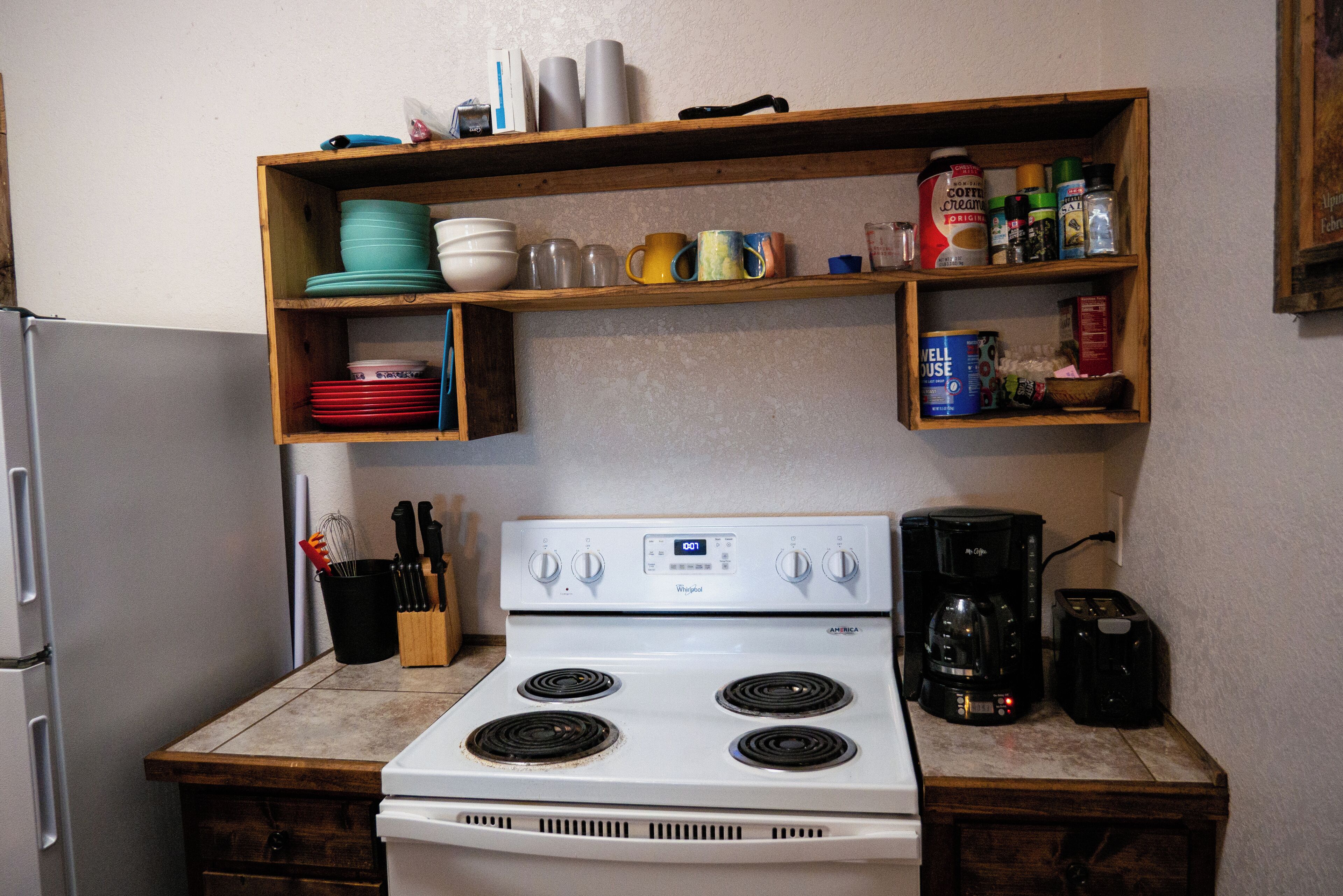 A kitchen with a white stove, a coffee maker, and shelves above holding dishes and spices.