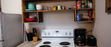 A kitchen with a white stove, a coffee maker, and shelves above holding dishes and spices.