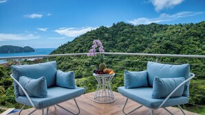 A balcony with two blue cushioned chairs, a small table with fruits and flowers, and a scenic view of a forested hillside and coastline. - Casa Colochos at Los Suenos Resort (Playa Herradura)