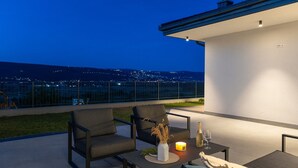 A balcony with two chairs, a table, a candle, and a bottle, overlooking a cityscape at dusk.