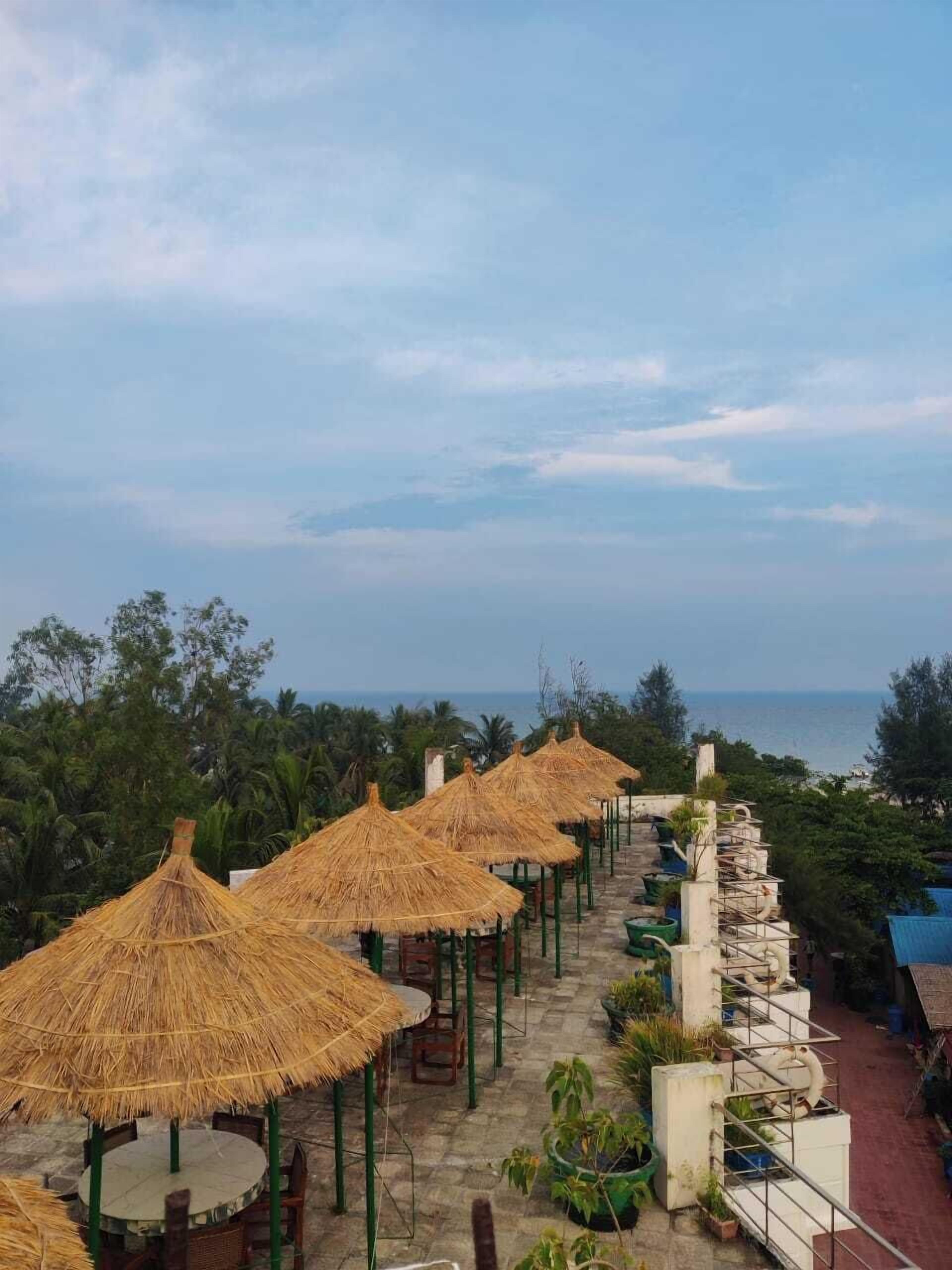 A terrace with thatched umbrellas and outdoor seating, overlooking a coastal area with palm trees and the sea.