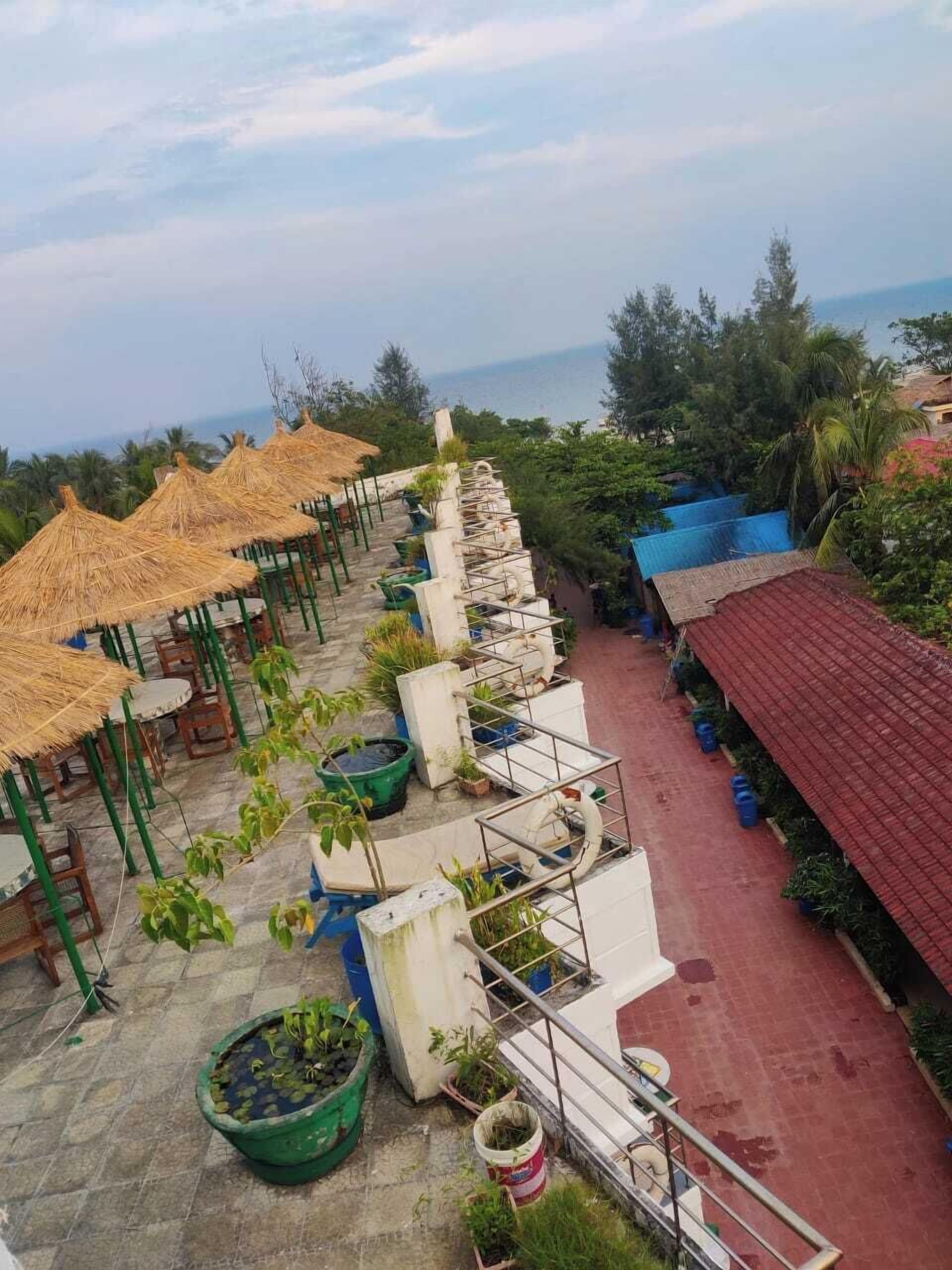 A rooftop terrace with thatched umbrellas, potted plants, and a red-roofed building.
