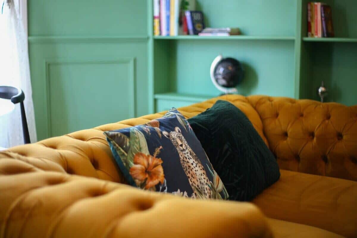 Un canapé jaune à motifs, agrémenté d’un coussin à fleurs et d’un coussin vert foncé, dans une pièce aux murs verts et où l’on aperçoit une bibliothèque à l’arrière-plan.