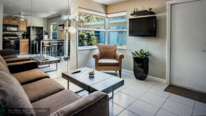 Una sala de estar moderna con un sofá, una mesa de centro y un sillón. Hay una zona de cocina con armarios y un refrigerador. Un televisor está montado en la pared, y una ventana ofrece vistas a la vegetación exterior.