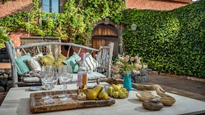 Een gezellig buiten zitgedeelte met een houten bankje, een tafel gedekt met wijnglazen, fruit en een bloevas.