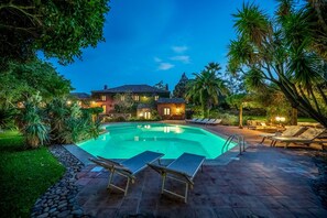 A swimming pool with lounge chairs, surrounded by a landscaped garden and a house with outdoor lighting.