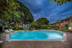 A rectangular swimming pool with a tiled deck, surrounded by lush greenery and a stone wall.