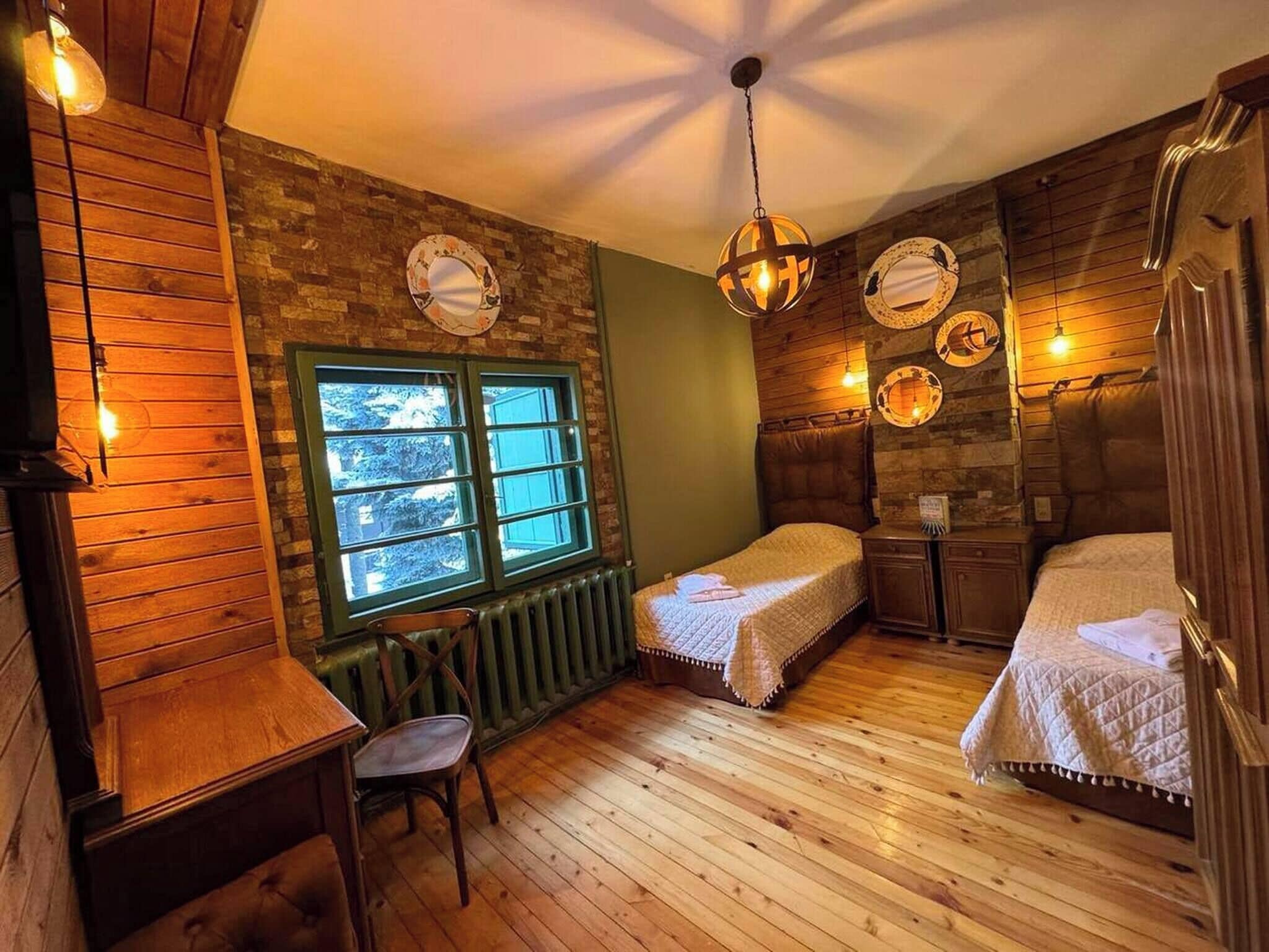 A cozy room with wooden floors, a green wall, a window with a view, two beds, a small table, and a chair.