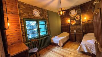 A cozy room with wooden floors, a green wall, a window with a view, two beds, a small table, and a chair.