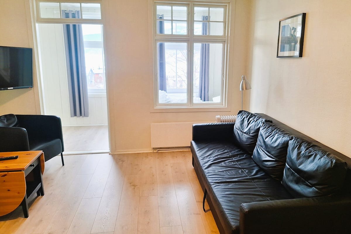 Een woonkamer met een zwarte leren bank, een houten salontafel en een flatscreen televisie. Er is een groot raam met gordijnen, een radiator en een ingelijst schilderij aan de muur.