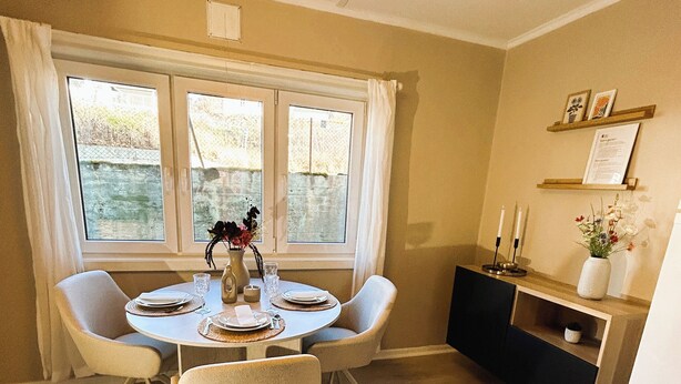 Un coin repas avec une table ronde, quatre chaises, un vase de fleurs et une fenêtre avec des rideaux.