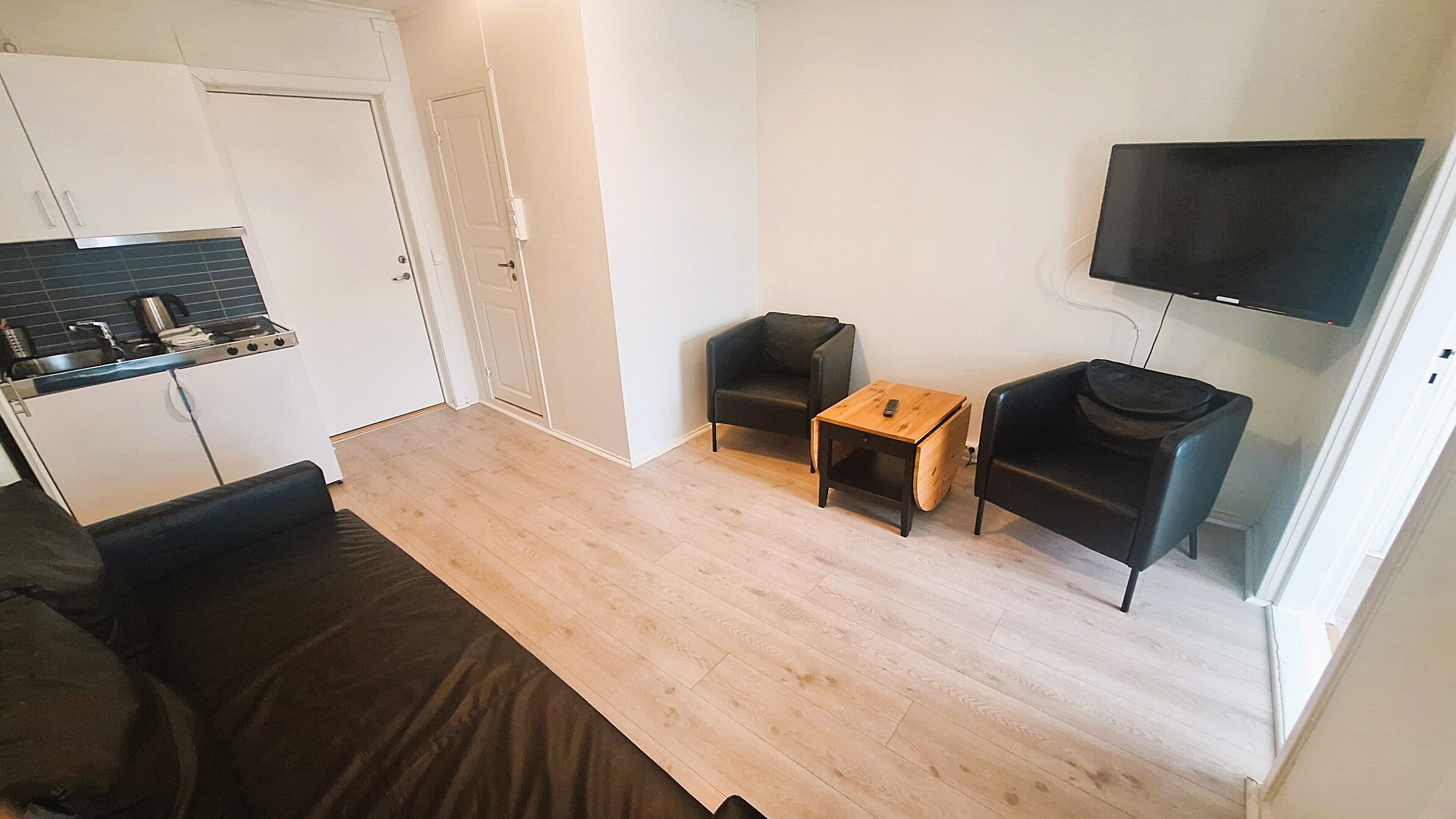 Ein kompakter Wohnraum mit Küchenzeile, einer Couch, einem kleinen Tisch, einem Stuhl und einem an der Wand befestigten Fernseher.