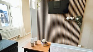 Un salon moderne avec un mur en lambris de bois, un téléviseur fixé au mur, un canapé, une table basse et une fenêtre donnant sur un immeuble.