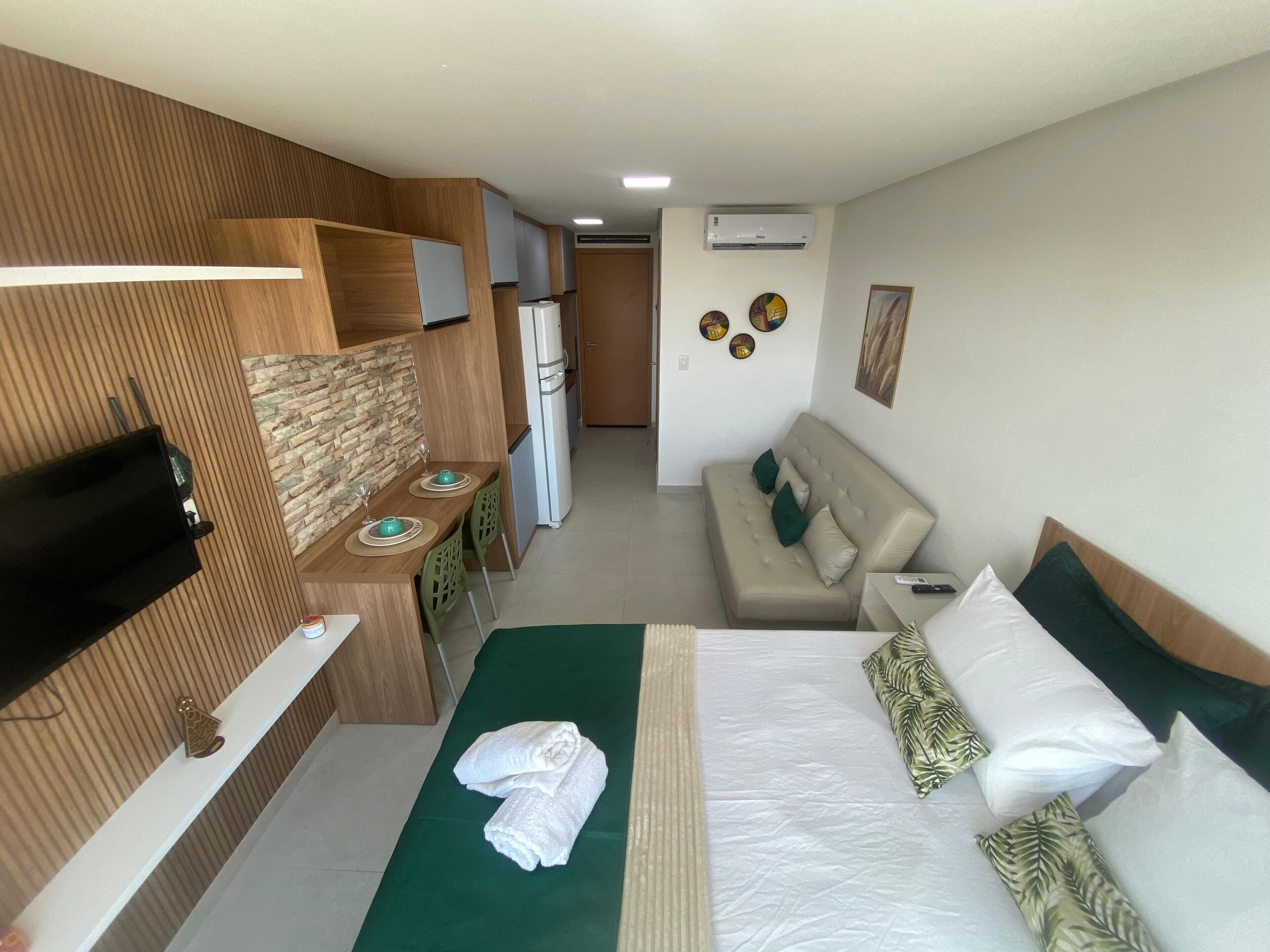 A hotel room with a bed, a sofa, a dining area with a table and chairs, a TV, and a refrigerator.