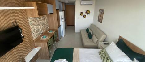 A hotel room with a bed, a sofa, a dining area with a table and chairs, a TV, and a refrigerator.