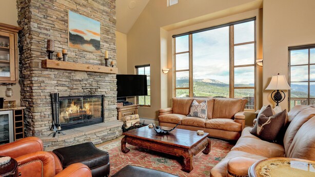 A spacious living room with a stone fireplace, leather sofas, and large windows offering a mountain view. - Luxurious Moonlight Basin Chalet! Stunning Views with Private Deck with Grill an (Big Sky)