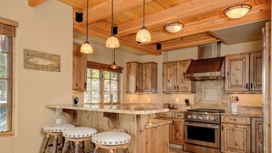 A rustic kitchen with wooden cabinets, a central island with stools, a stove, and pendant lighting. - Luxurious Moonlight Basin Chalet! Stunning Views with Private Deck with Grill an (Big Sky)