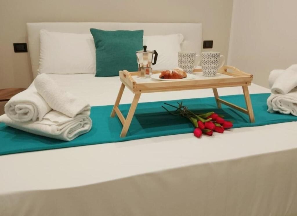 A neatly made bed with a teal bedspread, a wooden tray with a coffee pot, two patterned cups, and a croissant.