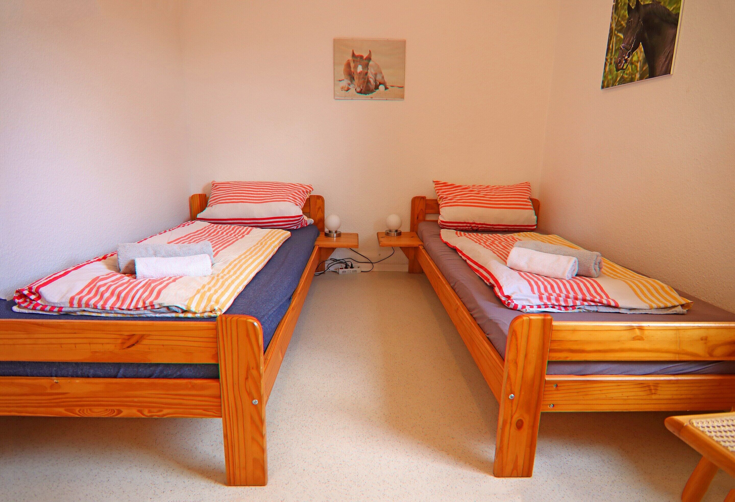 A room with two wooden beds, each with a pillow and a blanket. There is a small table between the beds, and a picture on the wall above the beds.
