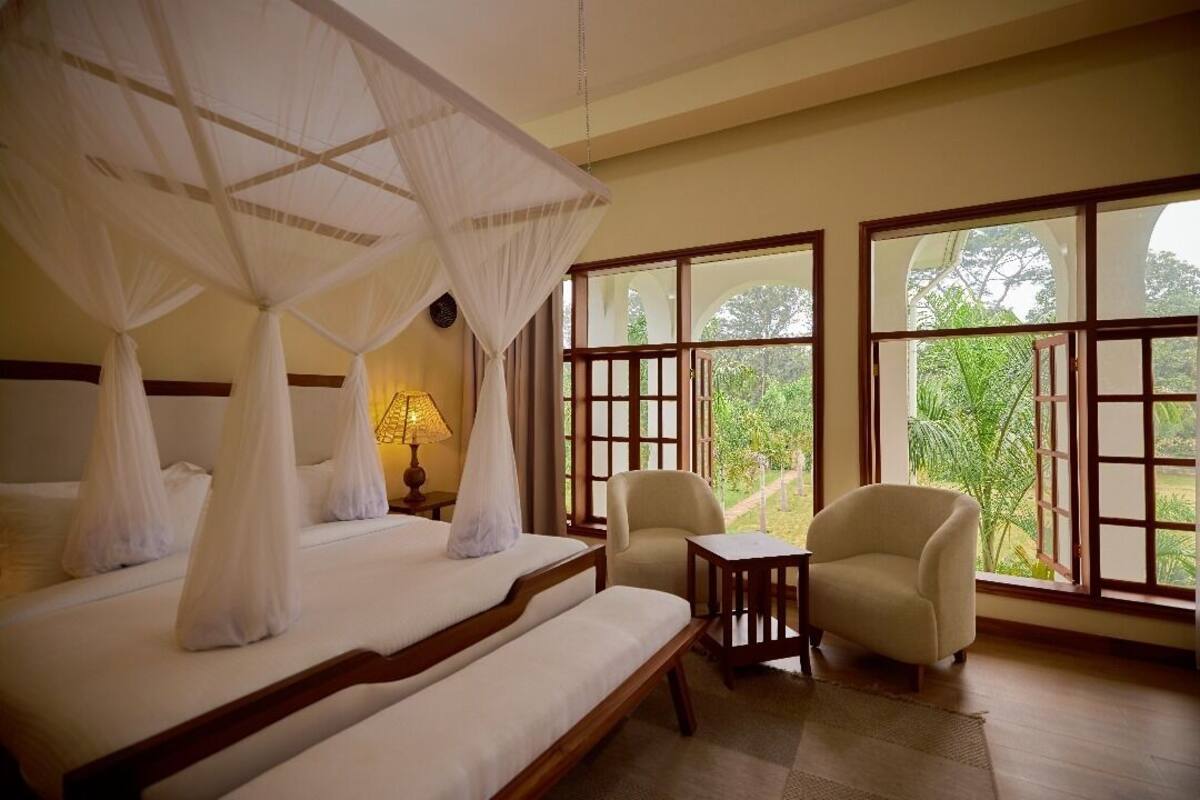 A bedroom with a bed, a chair, a small table, and large windows with a view of greenery.