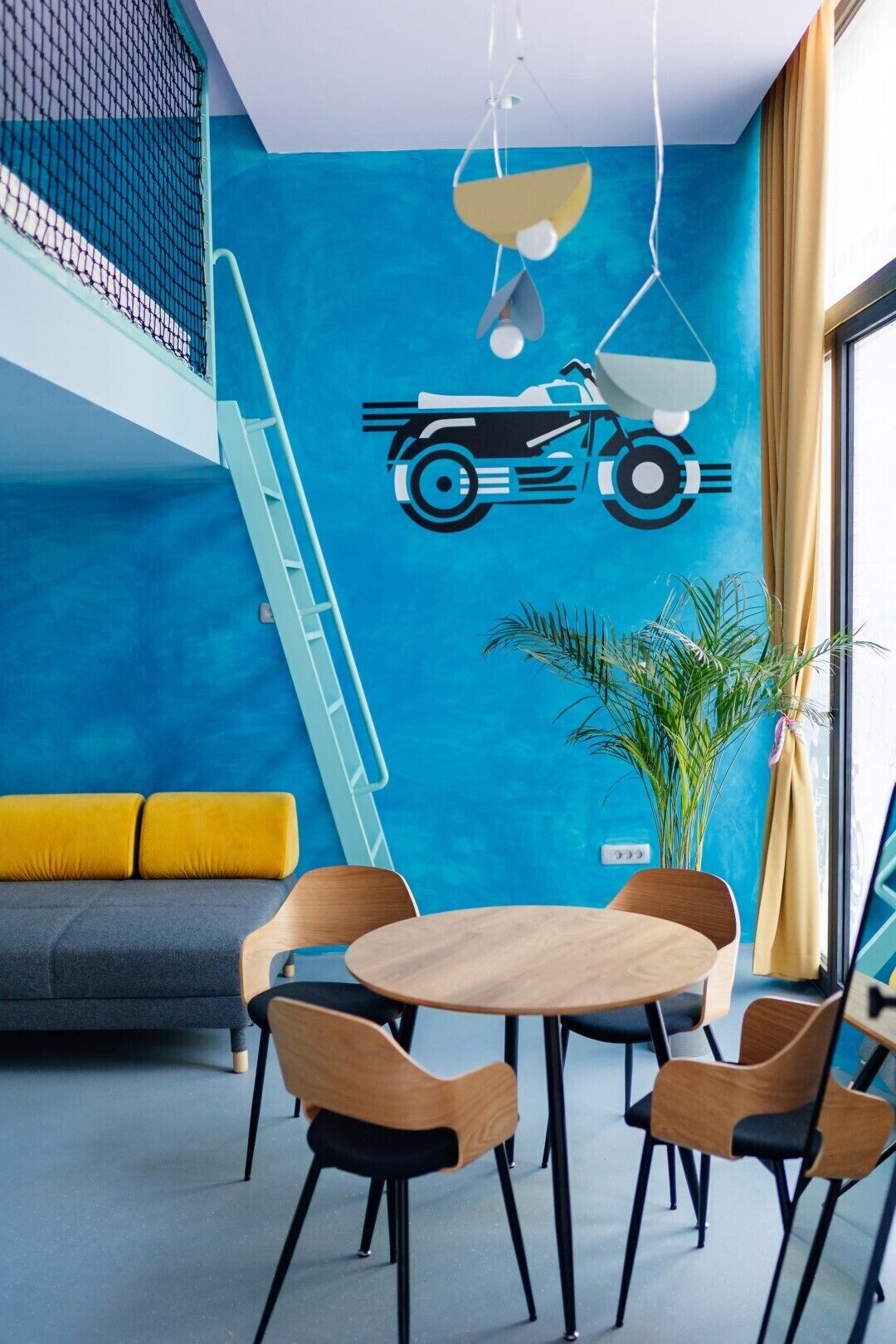 A modern room with a blue accent wall, a yellow sofa, a wooden dining table with chairs, and a motorcycle decal.