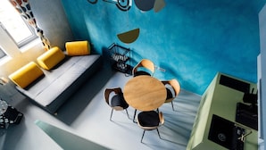 Quarto moderno com parede azul, sofá com almofadas amarelas, mesa redonda de madeira com cadeiras e uma pequena estante preta.