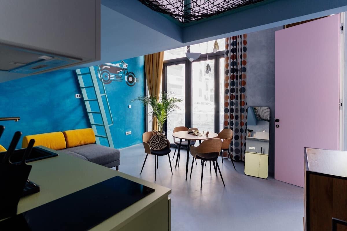 A modern room with a blue accent wall, a dining area with a round table and chairs, a sofa with yellow cushions, and a large window with curtains.