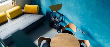 A room with a blue wall, a sofa with yellow cushions, a wooden table with chairs, and a black shelf with glasses.