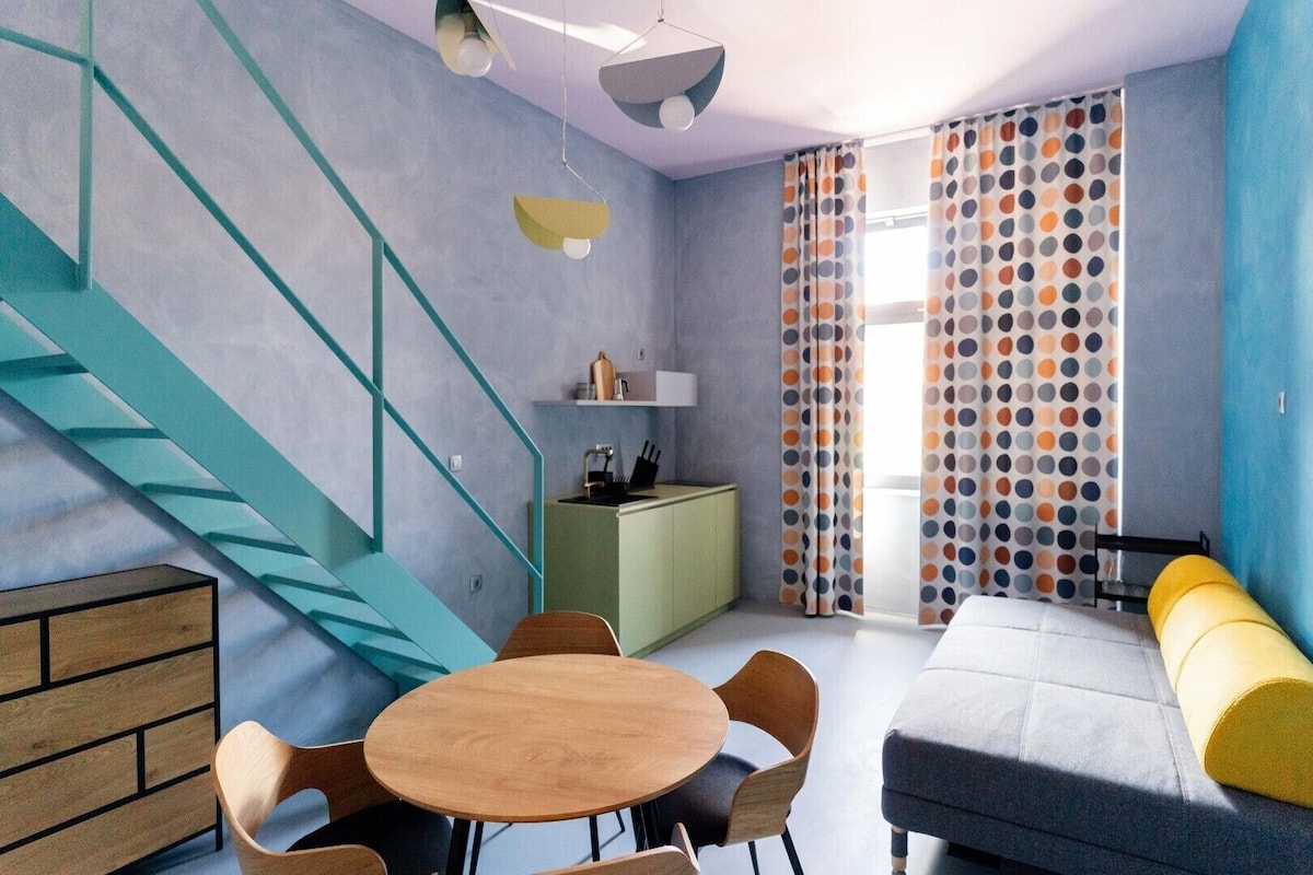 A modern room with a staircase, a dining area, a sofa, and a kitchen area.