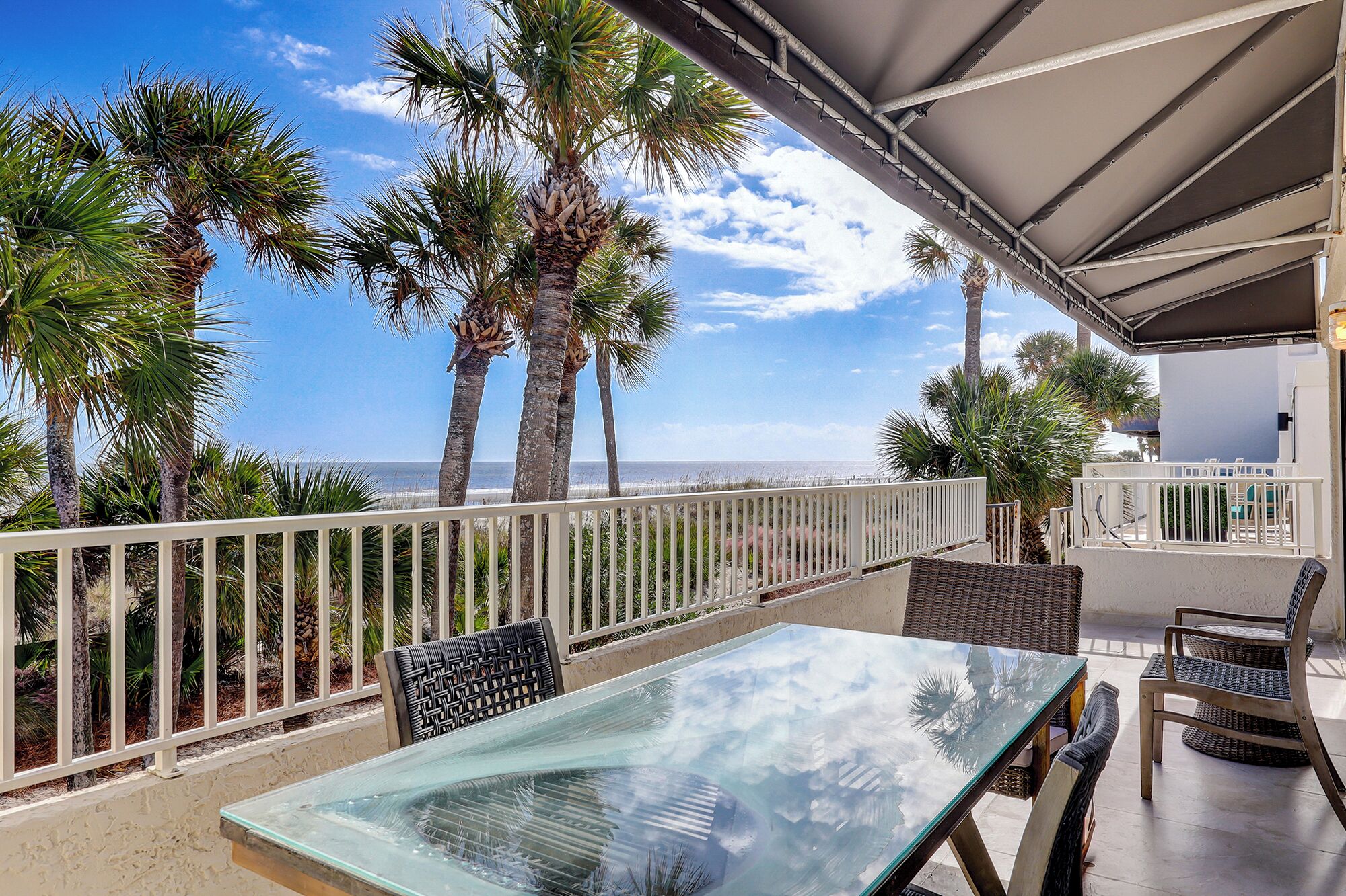 A balcony with a glass table and chairs, overlooking a beach with palm trees and the ocean.