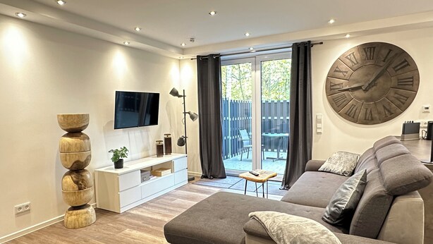 A modern living room with a large sofa, a TV mounted on the wall, a wooden floor, and a large clock. - Elegant new building between Hamburg & Lübeck, Baltic Sea with parking lot in Reinfeld (Reinfeld)