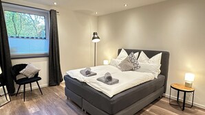 A modern bedroom with a large bed, bedside lamps, a chair, and a window with curtains. - Elegant new building between Hamburg & Lübeck, Baltic Sea with parking lot in Reinfeld (Reinfeld)
