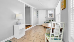 Una habitación con pared blanca, suelo de madera, una mesa de madera con sillas, una lámpara y una puerta que da a otra habitación.