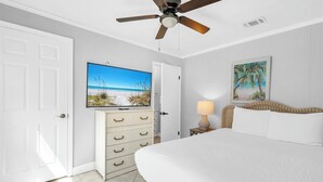 Un dormitorio con una cama, ventilador de techo, televisor, cómoda, lámpara y un cuadro.