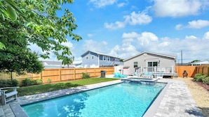 Una piscina rectangular con un deck de madera y sillas de descanso, rodeada por un cerco de madera y una casa al fondo.