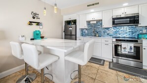 A modern kitchen with white cabinets, a stainless steel refrigerator, a microwave, and an oven. There is a bar area with white stools and a small island with a sink. - Spectacular Ocean View STEPS from the Beach and Pool! Third Floor Studio! (New Smyrna Beach)