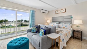 A modern bedroom with a large bed, a sofa, and a small table. There is a balcony with a view of the street and palm trees. - Spectacular Ocean View STEPS from the Beach and Pool! Third Floor Studio! (New Smyrna Beach)