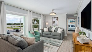 A modern living room with a large window, grey sofas, a teal armchair, and a wooden sideboard.
