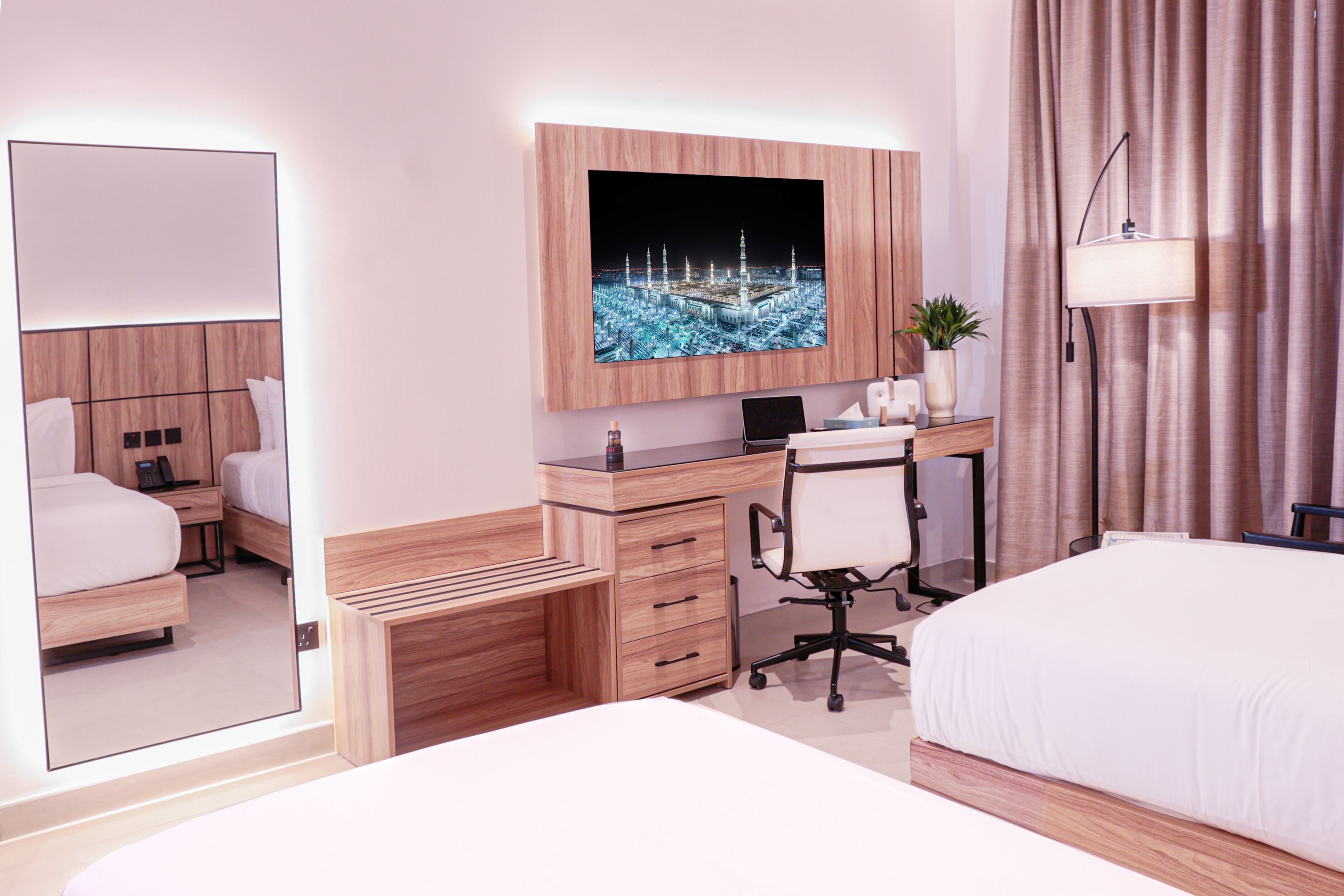 A modern hotel room with a large bed, a desk with a computer, a chair, a wooden desk, a mirror, and a TV displaying a cityscape.