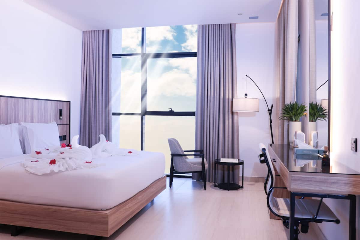 A modern hotel room with a large bed, a desk, and a chair. There is a window with curtains, a lamp, and a plant.