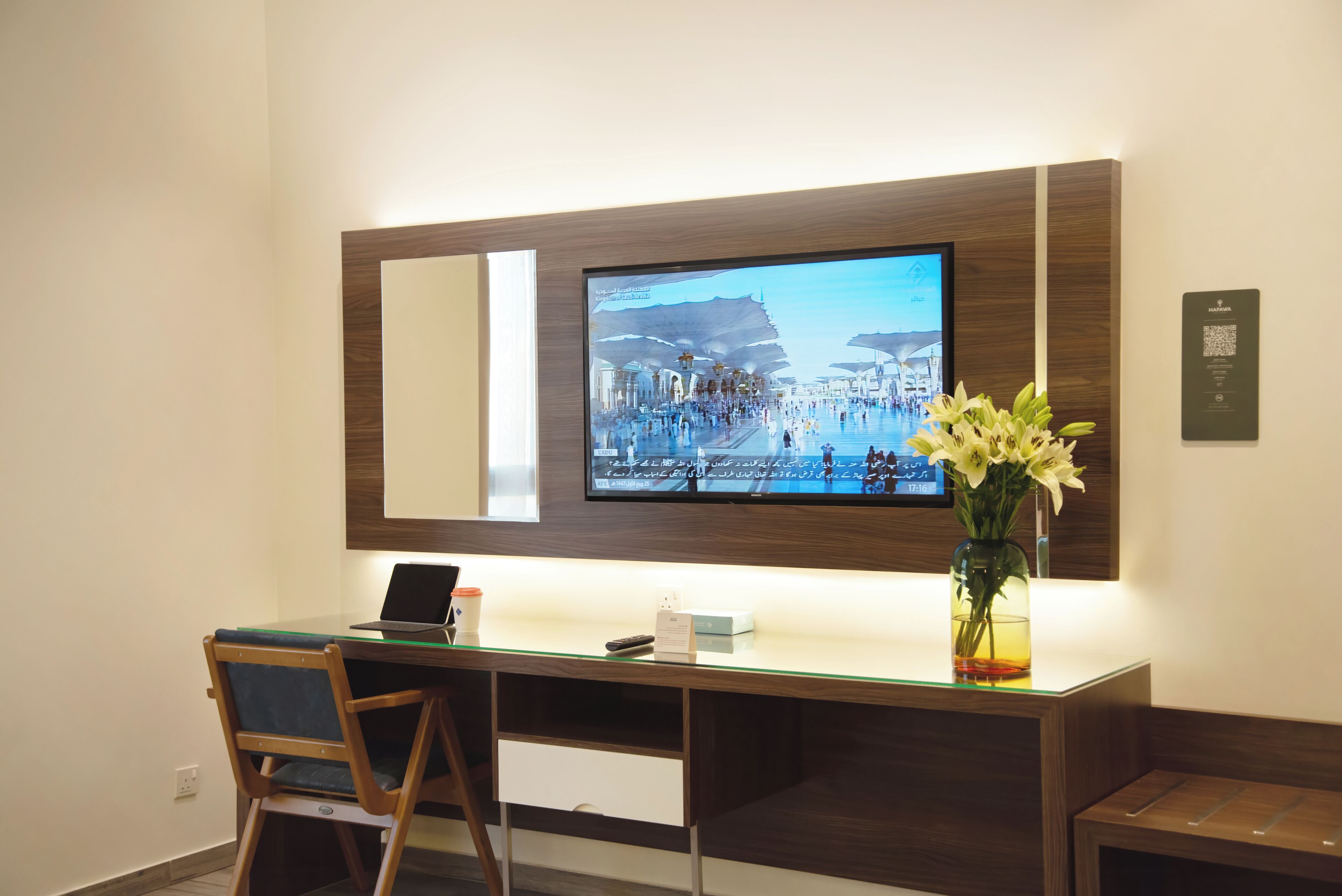 Ein modernes Hotelzimmer mit großem Flachbildfernseher an der Wand, einem Glastisch mit Laptop, einem Stuhl, einer Vase mit Blumen und einem kleinen Tisch mit Fernbedienung.