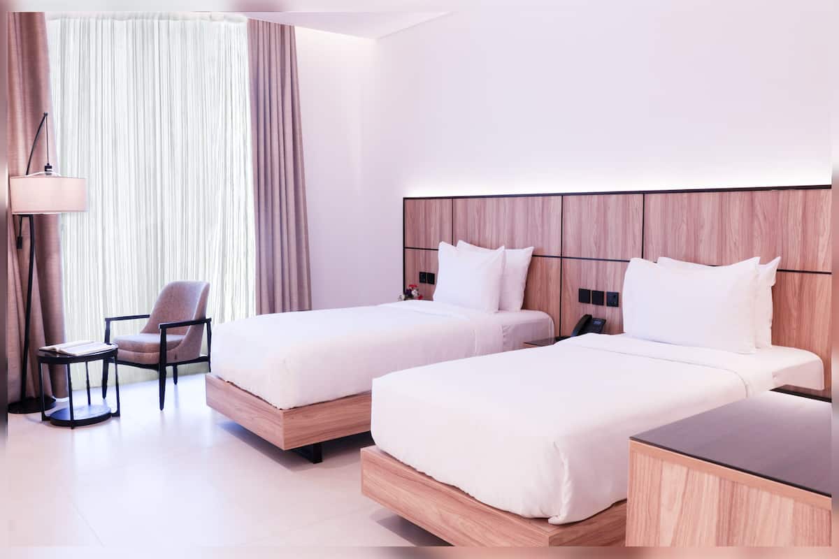 A hotel room with two beds, a chair, a small table, and a lamp.