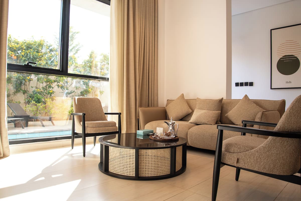 A modern living room with a round coffee table, two armchairs, and a sofa. A large window with curtains provides a view of an outdoor pool and greenery.