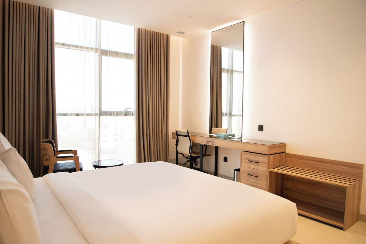 A hotel room with a large bed, a desk, a chair, a mirror, and a view of the city through large windows.