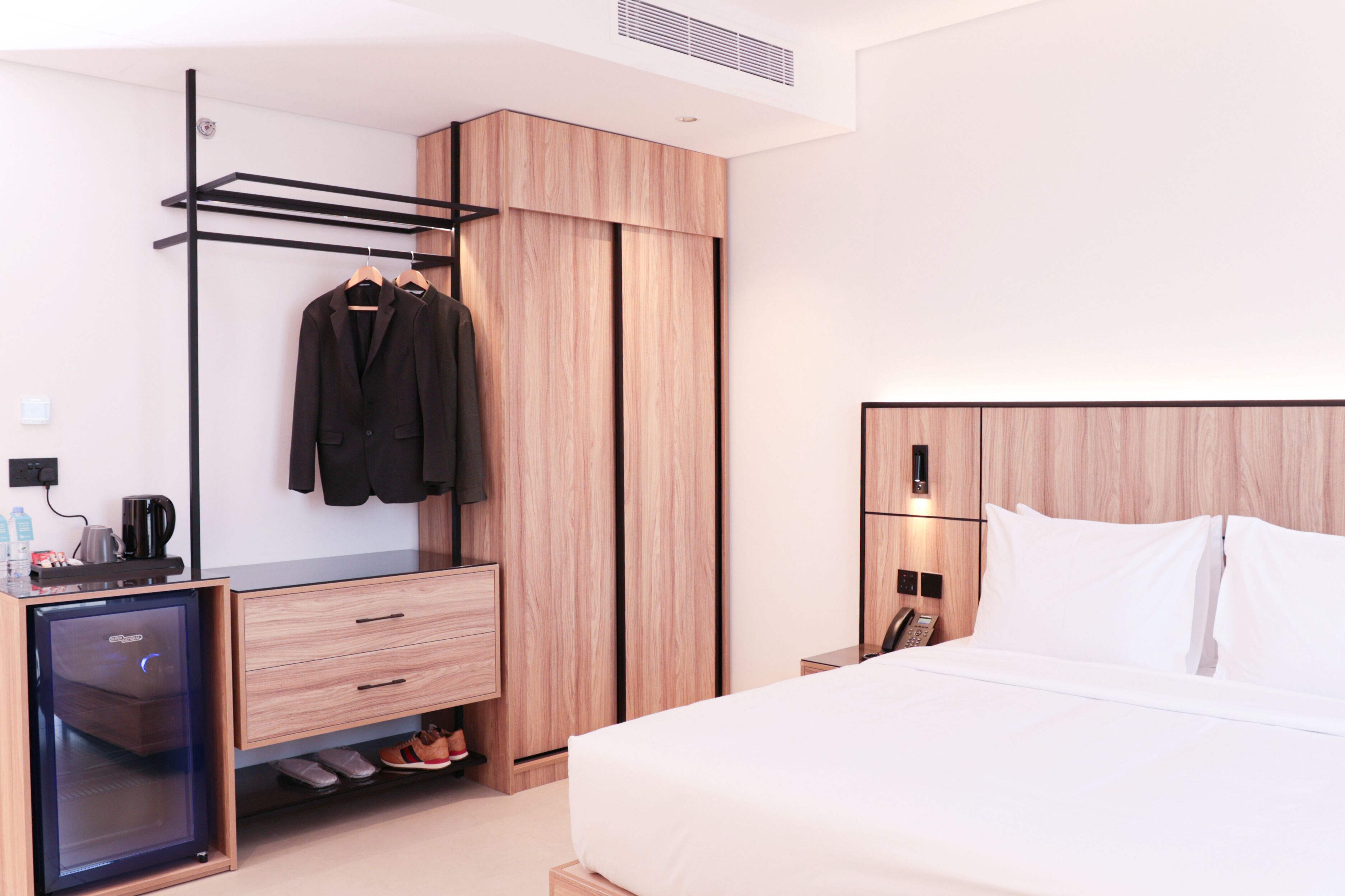 Una habitación de hotel moderna con una cama, un armario, un pequeño escritorio con refrigerador, un zapatero y una mesita de noche con teléfono.